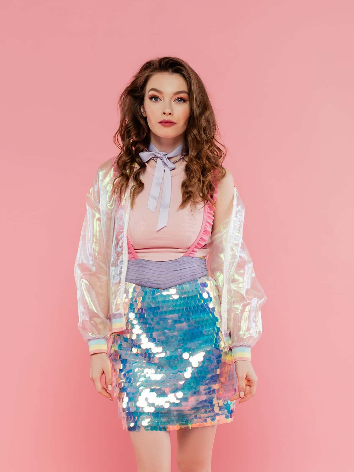 beautiful woman with wavy hair posing like a doll on pink background, conceptual photography, girly outfit, model in skirt with sequins and transparent jacket looking at camera